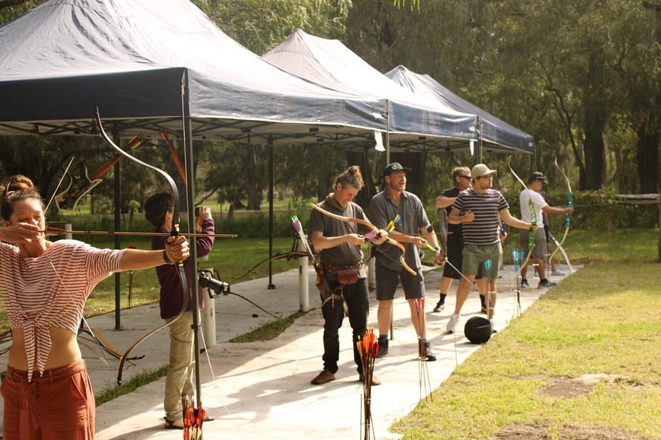 A group of archers lined up outdoors under shaded canopies, each drawing their traditional bows in unison, creating a strong sense of teamwork and focus. The setting features lush greenery and a soft shooting area