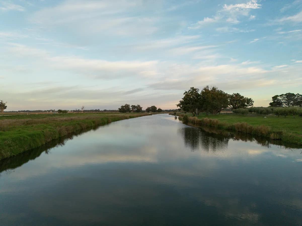 A river running between green paddocks, seen from the air, with trees reflected in the still water under a wide sky.