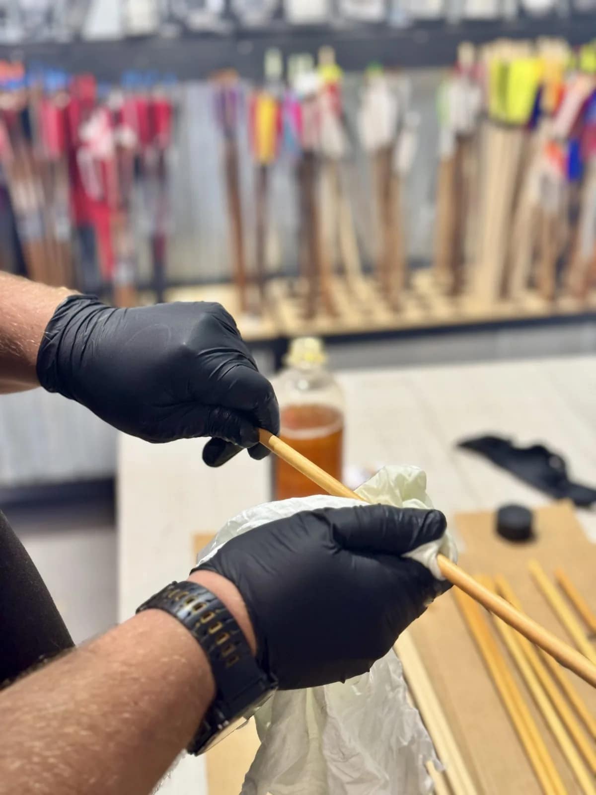 Two gloved hands wiping finish onto a bare wooden arrow shaft at a workbench, with a jar of amber stain, more bare shafts beside it and racks of brightly fletched arrows behind
