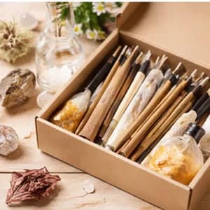 A discovery box of incense: mixed sticks wrapped in paper, packed upright in a kraft carton with dried botanicals beside it.