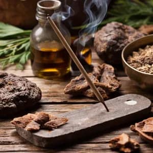 Grounding incense: a smoking stick on a weathered wooden holder beside tree bark, resin and a bottle of oil.