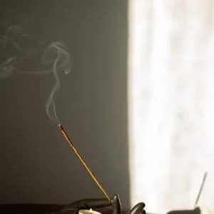 An incense stick burning in a dark cast holder, smoke rising against a sunlit wall.