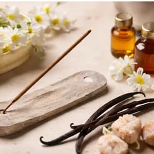 Sweet and warm incense: a stick on a pale wooden holder beside vanilla pods, raw sugar and amber oils.